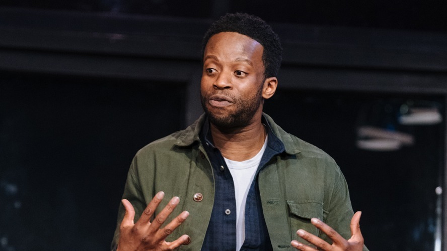 Clifford Samuel as Sam, wearing a dark coat, stands in a dimly lit, atmospheric setting.