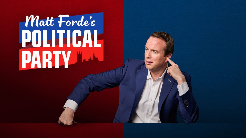 Matt Forde, in a blue suit, gestures thoughtfully against a vibrant red and blue background.