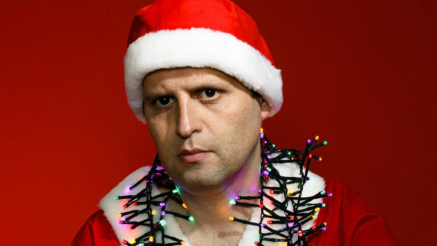 Adam Kay stands on stage, speaking into a microphone, with a spotlight illuminating him.