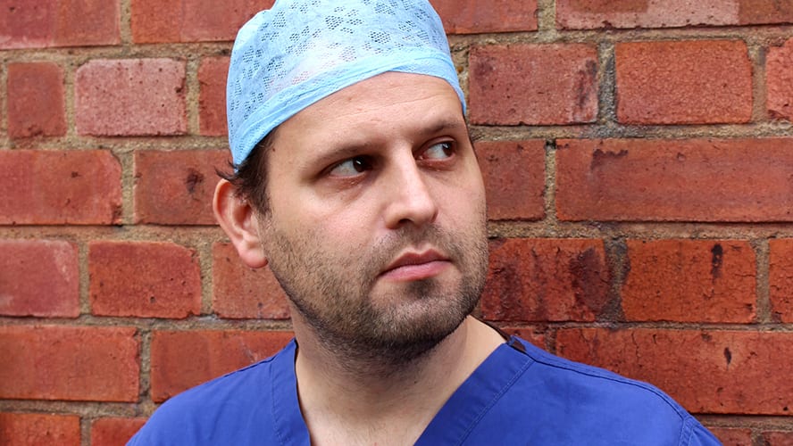 Adam Kay standing on stage, speaking into a microphone, with an audience in the background.