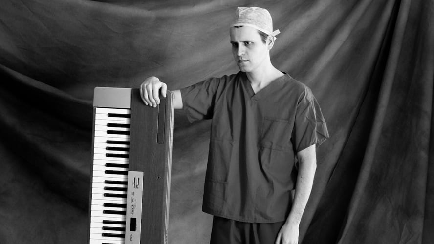 Adam Kay stands on stage, speaking into a microphone, with an audience visible in the background.