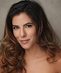 Aimie Atkinson smiling, wearing a stylish black dress with elegant makeup, against a neutral background.
