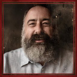 A bearded man smiles warmly, wearing a light grey shirt against a dark, textured background.