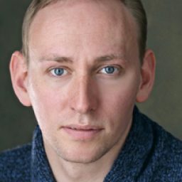 Alex Frost smiling, wearing a blue shirt, with short brown hair and glasses, against a neutral background.