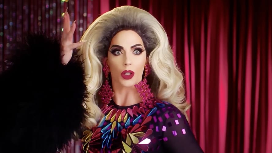 Alyssa Edwards smiling, wearing a vibrant outfit with dramatic makeup, against a colourful background.