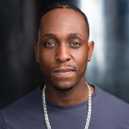 Ashley D Gayle, a Black man with short hair, wearing a dark shirt, looking directly at the camera.
