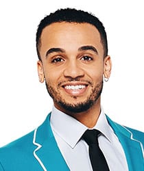 Aston Merrygold smiling, wearing a stylish outfit, with a blurred background of lights.