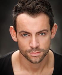 Ben Forster smiling, wearing a dark suit and white shirt, with short brown hair.