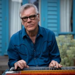 BJ Cole, a musician, poses with a steel guitar, smiling against a neutral background.