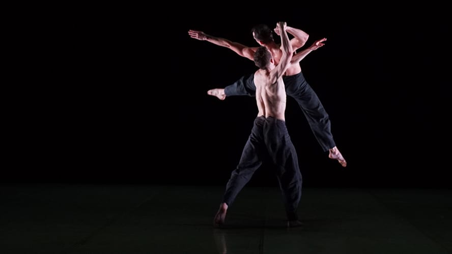 Two male dancers in black outfits performing a dynamic lift on stage.