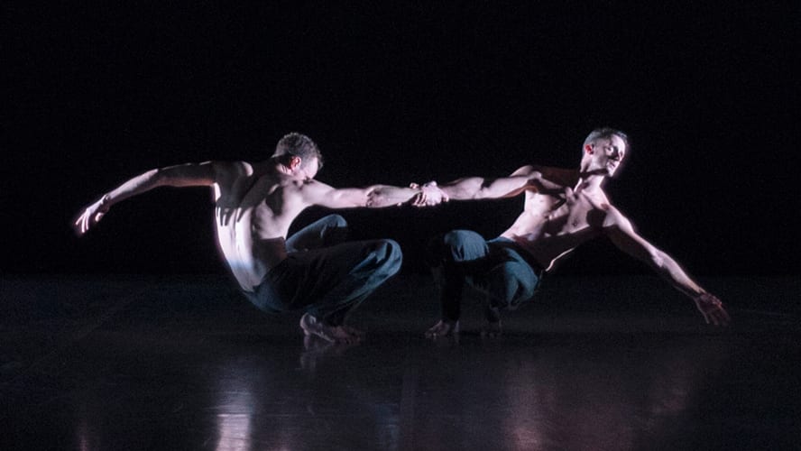 Two male dancers in black attire performing a dynamic lift on a stage with dramatic lighting.