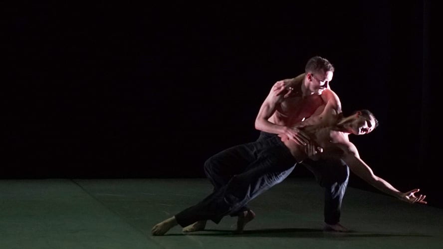 Two male dancers in black attire performing a dynamic lift on stage.