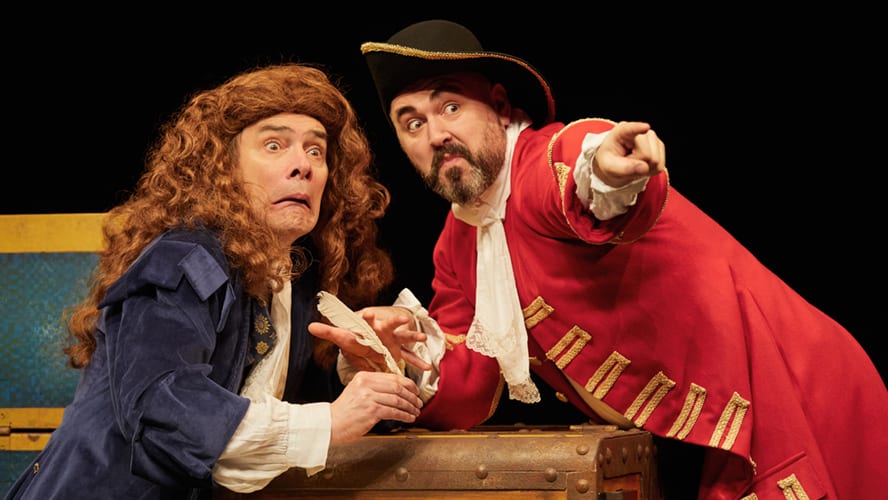 A vibrant stage scene featuring actors in historical costumes performing in front of a large audience.