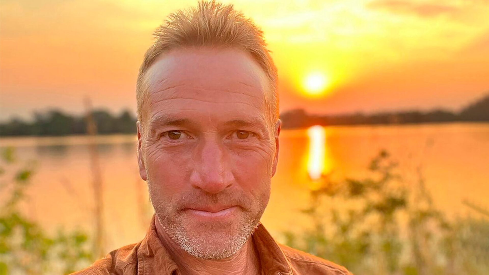 Ben Fogle smiling outdoors, wearing a casual jacket, with greenery and trees in the background.