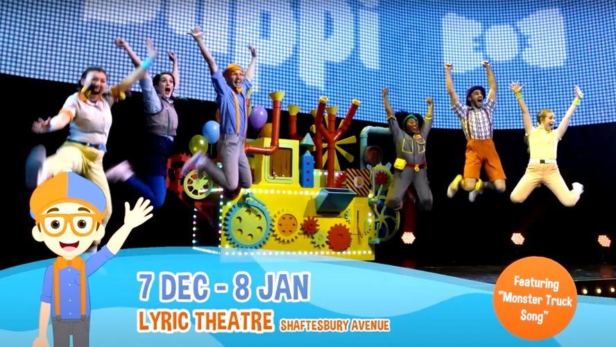 Blippi wearing an orange beanie and blue shirt, smiling and standing in a colourful outdoor setting.