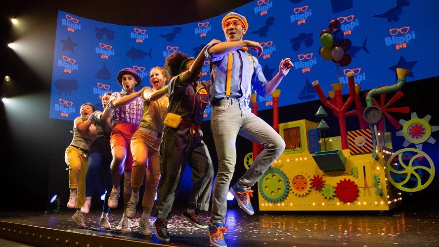 Blippi in a vibrant costume, performing energetically on stage with colourful props and a lively backdrop.