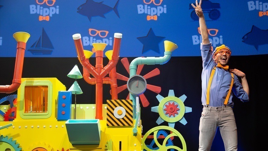 Blippi performing on stage, surrounded by colourful set pieces and enthusiastic young audience members.