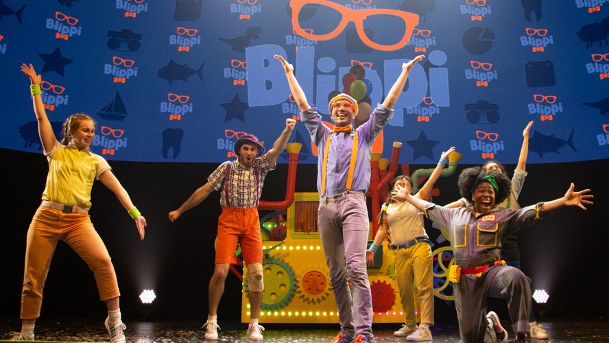 Blippi performing on stage, surrounded by colourful set pieces and enthusiastic young audience members.
