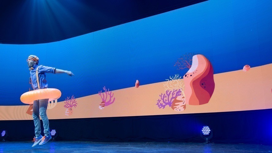 Blippi performing on stage, surrounded by colourful set pieces and enthusiastic young audience members.
