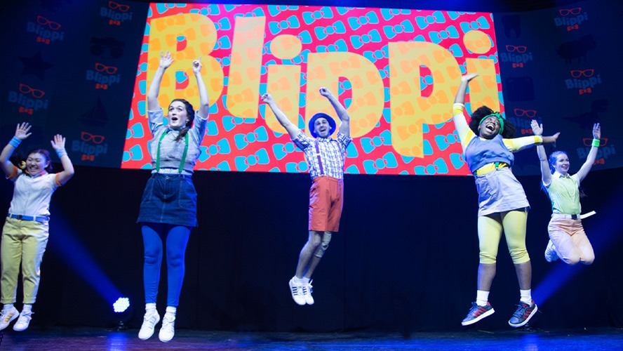 Blippi performing on stage, surrounded by colourful set pieces and enthusiastic young audience members.