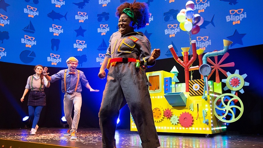 Blippi performing on stage, surrounded by colourful set pieces and enthusiastic young audience members.
