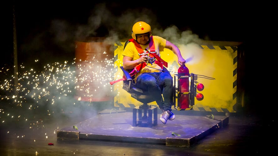 A lively stage scene with performers engaging the audience, surrounded by colourful props and lighting.