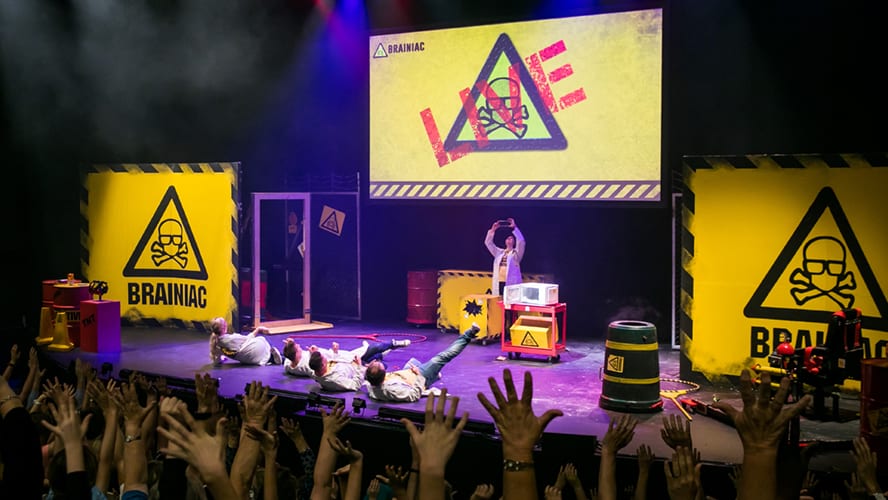 A stage set with colourful lights, featuring performers engaging in a science demonstration.