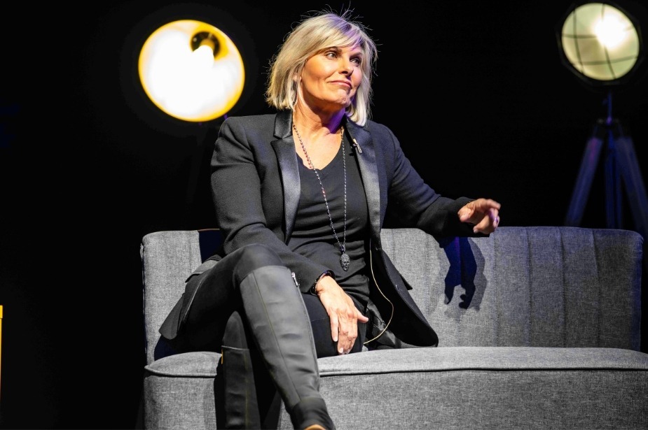 Captain Sandy smiling on stage, wearing a stylish outfit, with bright lights illuminating the background.