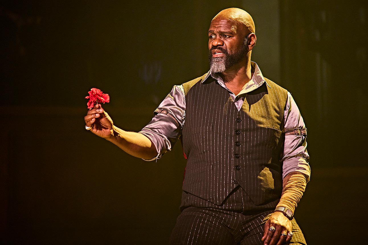 Chris Jarman as King Hades, holding a red flower, wearing a dark striped vest and shirt.