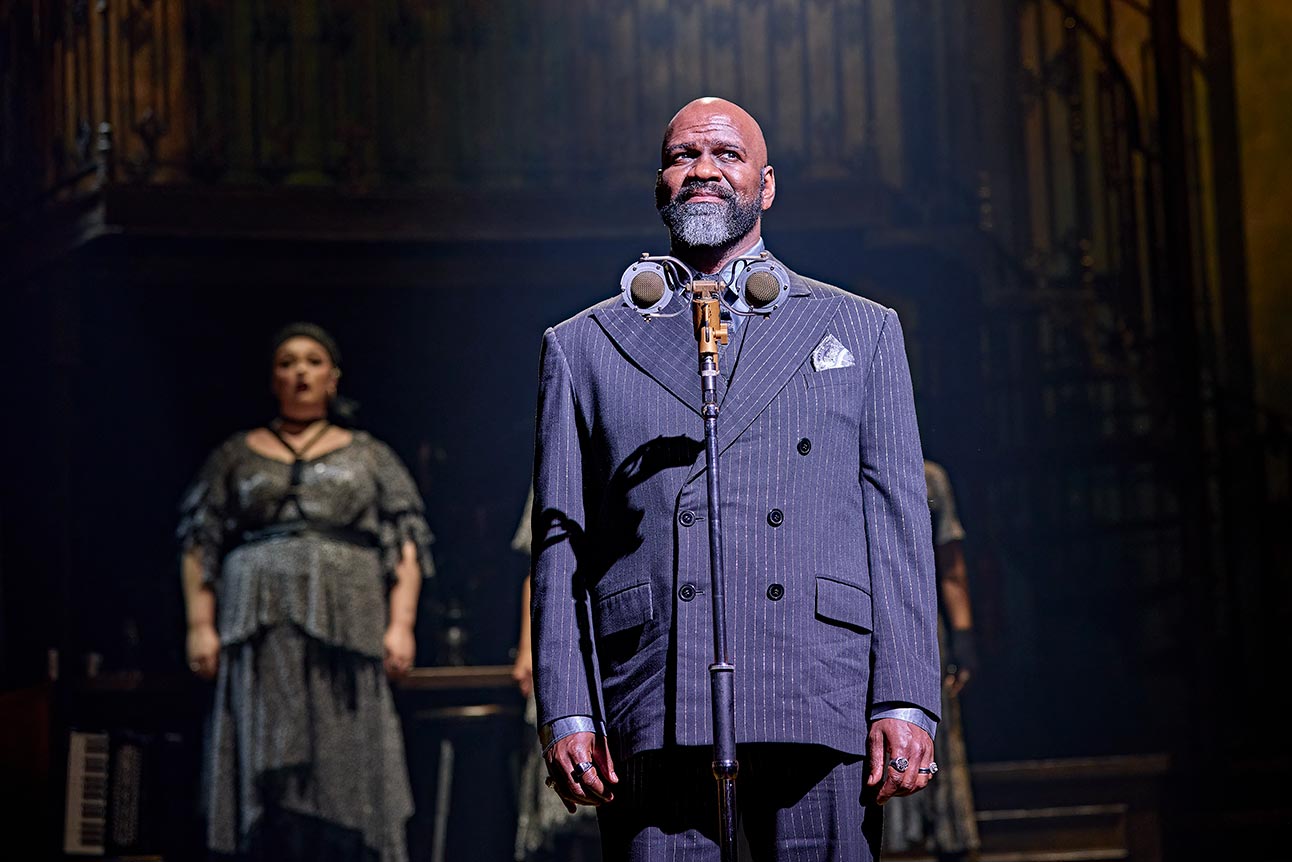 Chris Jarman as King Hades, wearing a pinstriped suit, stands confidently on stage.