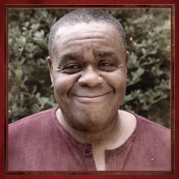 Clive Rowe smiling, wearing a maroon shirt, with a blurred leafy background.