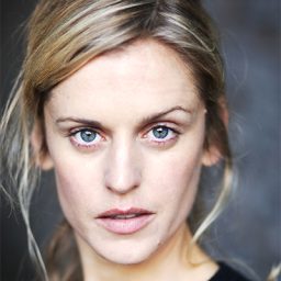 Denise Gough performing on stage, wearing a black outfit, with dramatic lighting and a focused expression.