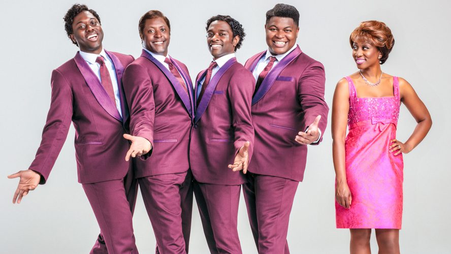 A group of four diverse performers in vibrant costumes, smiling and posing together on stage.