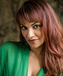 Danielle de Niese smiling, wearing an elegant dress, with her hair styled in soft waves.