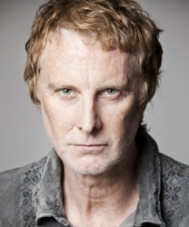 David Threlfall smiling, wearing a dark suit and tie, against a neutral background.