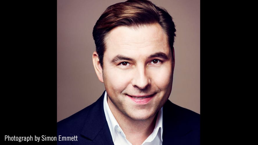 David Walliams smiling, wearing a dark suit, with a blurred background of soft lighting.