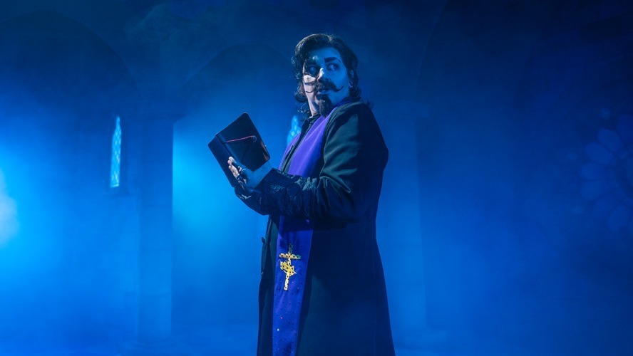 A performer in a vibrant costume poses dramatically on stage with theatrical lighting.