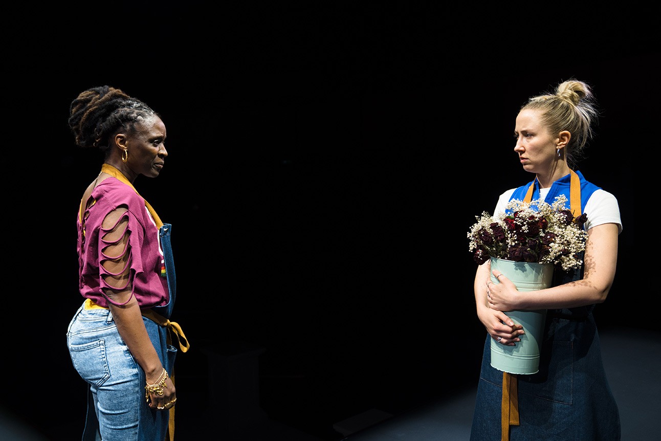 A theatrical scene featuring actors on stage, with dramatic lighting and a minimalist set design.