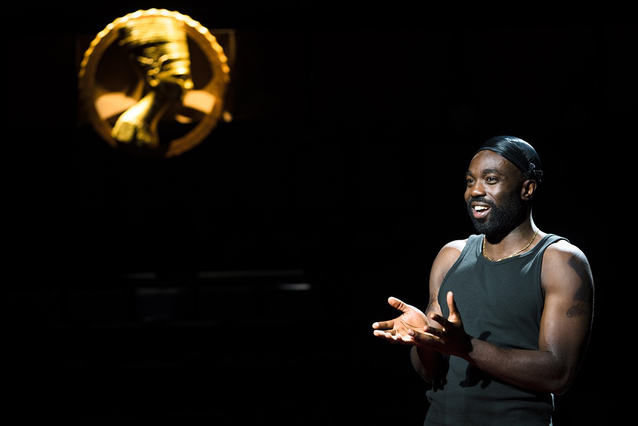 A dramatic stage scene featuring a male actor in a suit, illuminated by spotlight.