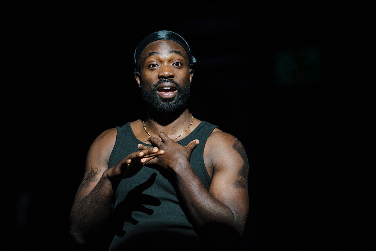 A theatrical scene featuring a male actor in a dramatic pose on stage, with dim lighting.