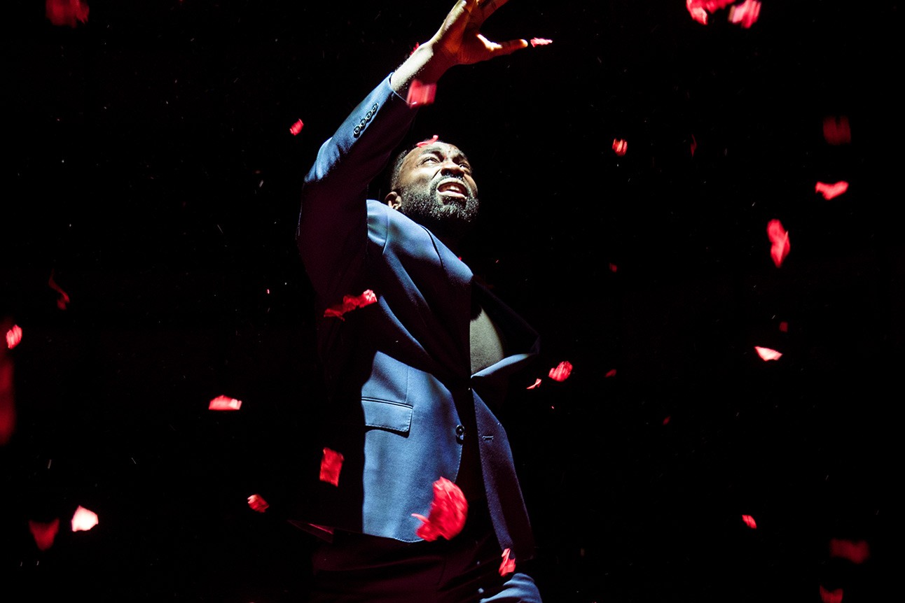 A theatrical scene featuring a male actor in a dramatic pose on a dimly lit stage.