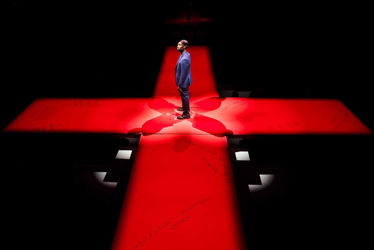 A theatrical scene featuring a man in a suit, standing on a stage with dramatic lighting.