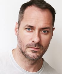 Declan Bennett smiling, wearing a black shirt, with short dark hair and a neutral background.