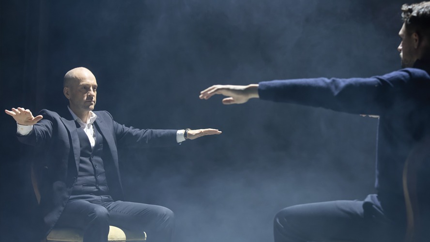 Derren Brown performing on stage, illuminated by dramatic lighting, with an engaged audience in the foreground.