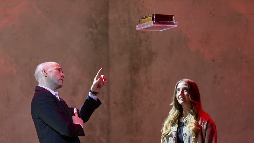Derren Brown performing on stage, spotlight illuminating him, with an audience visible in the background.