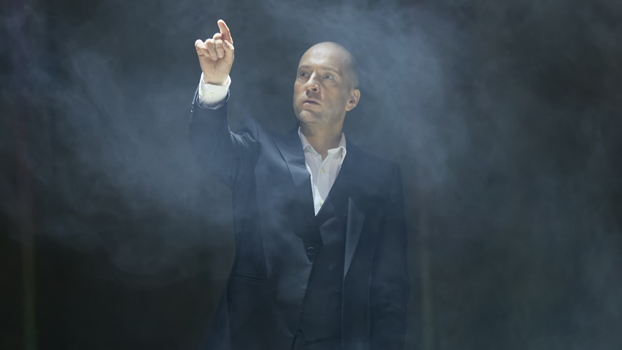 Derren Brown performing on stage, wearing a black suit, with dramatic lighting and an engaged audience.