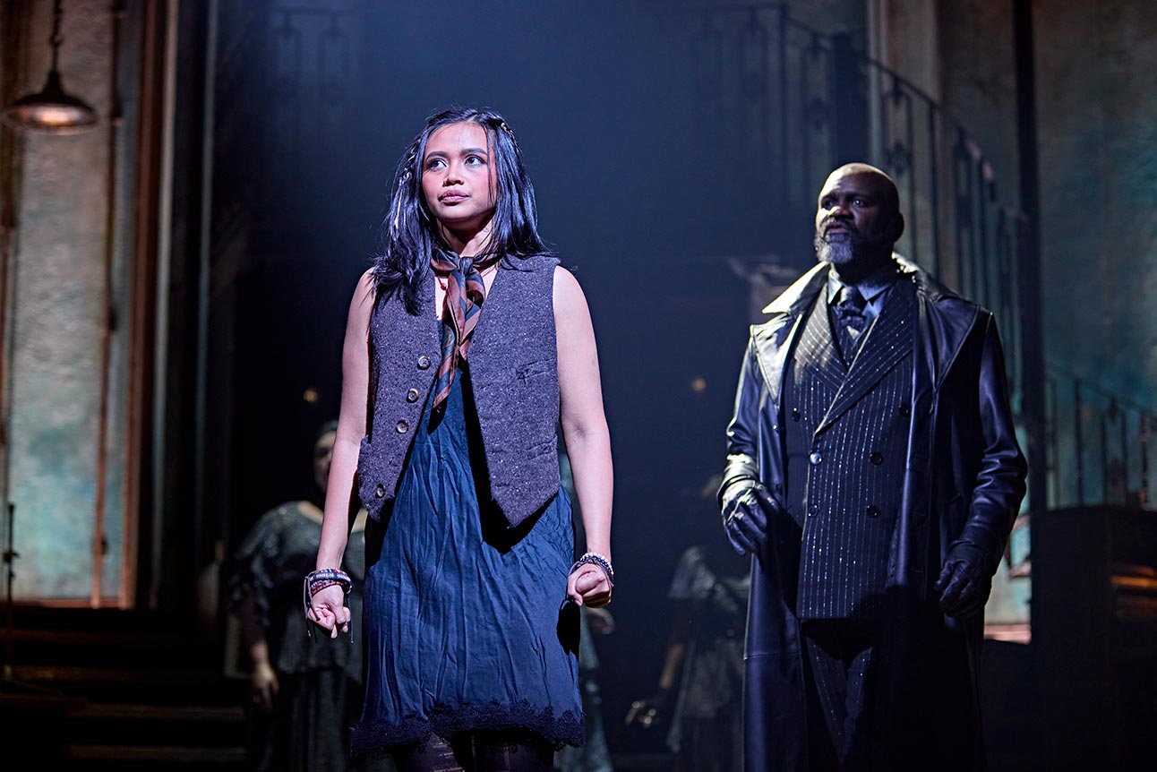 Desmonda Cathabel in a grey vest and dark blue dress stands confidently beside Chris Jarman in dim lighting.