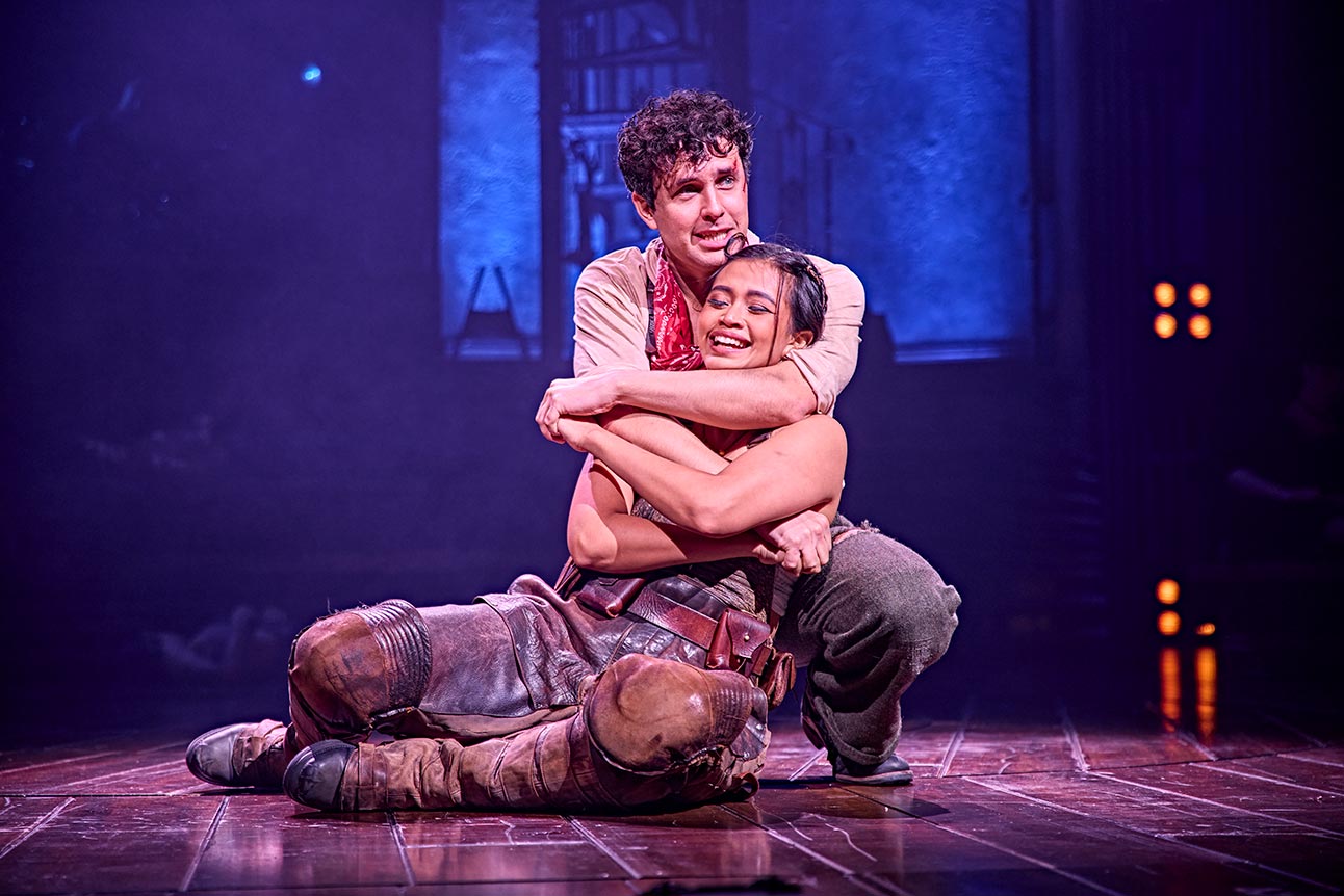 Desmonda Cathabel and Dylan Wood embrace on stage, surrounded by soft purple lighting and wooden flooring.