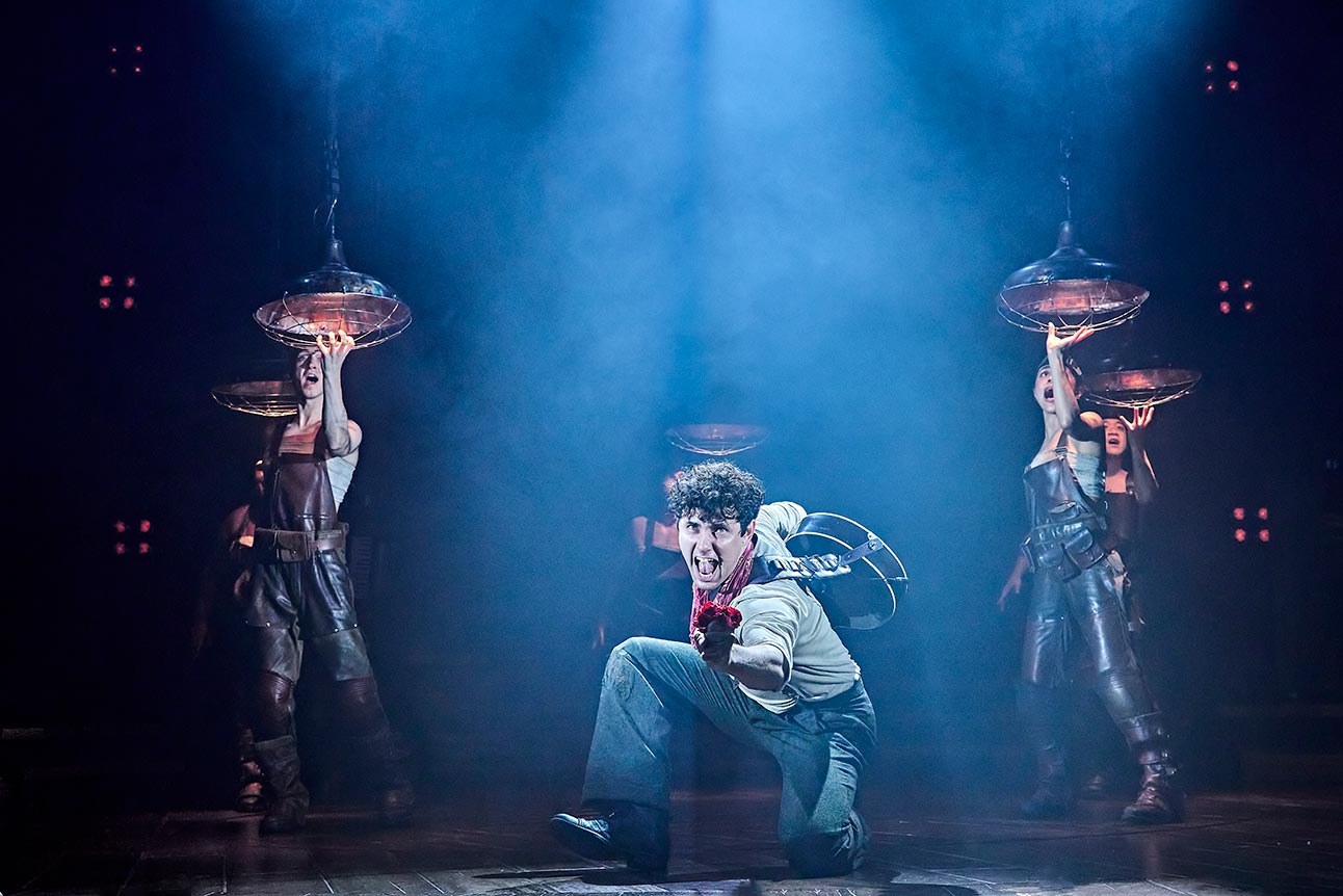 Dylan Wood as Orpheus, wearing a grey outfit, performs dramatically amidst smoky blue lighting.
