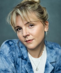 Eloise Davies smiling, wearing a blue blouse, with long brown hair and a neutral background.